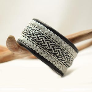 Könnte beinhalten: Ein schwarzes Lederarmband mit einem silbernen geflochtenen Design. Das Armband ist mit einem keltischen Knotenmuster gewebt.