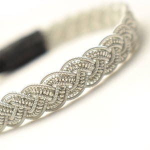 Puede incluir: Un brazalete trenzado plateado hecho de cuerdas de guitarra. El brazalete está tejido en un patrón complejo y tiene un acabado liso y pulido.