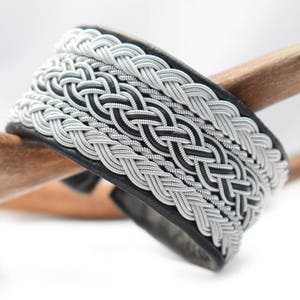 Könnte beinhalten: Ein schwarzes Lederarmband mit einem geflochtenen Silberdesign. Die Flechtung ist in einem komplexen Muster gewebt und hat eine glänzende Oberfläche.
