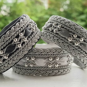 Könnte beinhalten: Nahaufnahme von drei silberfarbenen Armbändern mit aufwendigen geflochtenen Designs. Die Armbänder weisen dekorative silberne Perlen und blattförmige Akzente auf. Zwei Armbänder haben eine dunkelgraue Innenseite, das dritte eine hellgraue. Verschwommener grüner Hintergrund.