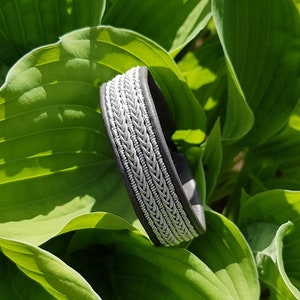 Könnte beinhalten: Ein graues Lederarmband mit einem silbernen geflochtenen Design. Das Armband liegt auf einem grünen Blatt-Hintergrund.