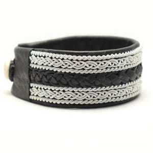 Könnte beinhalten: Schwarzes Lederarmband mit silbernem geflochtenem Besatz. Das Armband hat ein gewebtes Design und eine silberne Schließe.