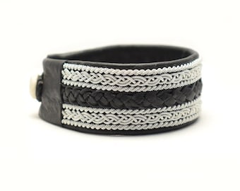 May include: Black leather bracelet with silver braided trim. The bracelet has a woven design and a silver buckle.