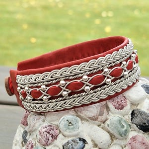 Könnte beinhalten: Ein rotes Armband mit aufwendigen Silberdrahtdetails und kleinen silbernen Perlen. Das Armband hat ein geflochtenes Design und rote ovale Akzente. Das Armband liegt auf einer Steinoberfläche.
