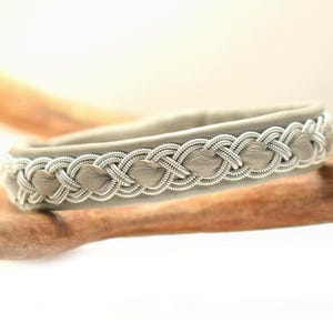 Könnte beinhalten: Ein graues Lederarmband mit silbernem Zopfmuster. Das Armband hat ein gewebtes Muster an den Rändern, wodurch ein strukturiertes und detailliertes Aussehen entsteht. Das Armband wird auf einer Holzoberfläche präsentiert.