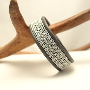 Könnte beinhalten: Ein graues Lederarmband mit einem geflochtenen Silberdesign. Das Armband liegt auf einem Geweih aus Holz.