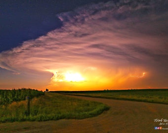 May include: A dramatic sunset with a large, dark cloud in the sky. Lightning strikes in the distance, illuminating the clouds with a bright orange glow. A dirt road leads into the distance, with a field of tall grass on either side.