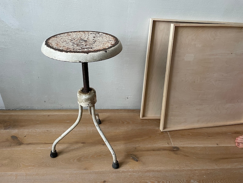 May include: White metal stool with a round, rusty top and three legs. The stool is on a wooden floor.