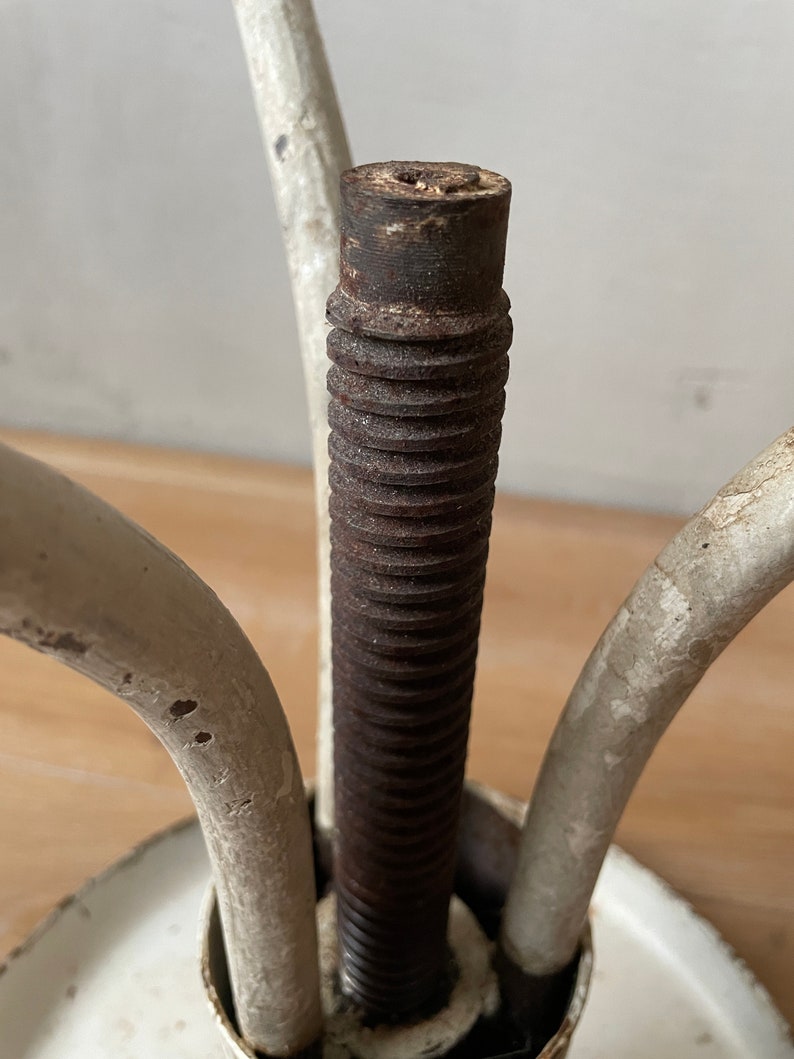 May include: Close-up of a white metal stool with a threaded metal rod and three legs. The rod is rusty and the legs are slightly chipped.