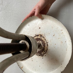 May include: White metal stool with a round base and three legs. The base is slightly rusted and the legs are adjustable.