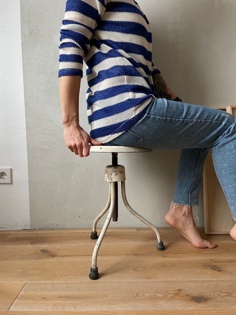 May include: White metal adjustable stool with three legs and a round seat. The stool is made of metal and has a white finish. The stool is adjustable in height.