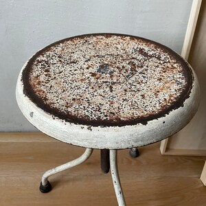 May include: White metal stool with a round, rusty top and three legs. The stool is slightly worn and has a vintage look.