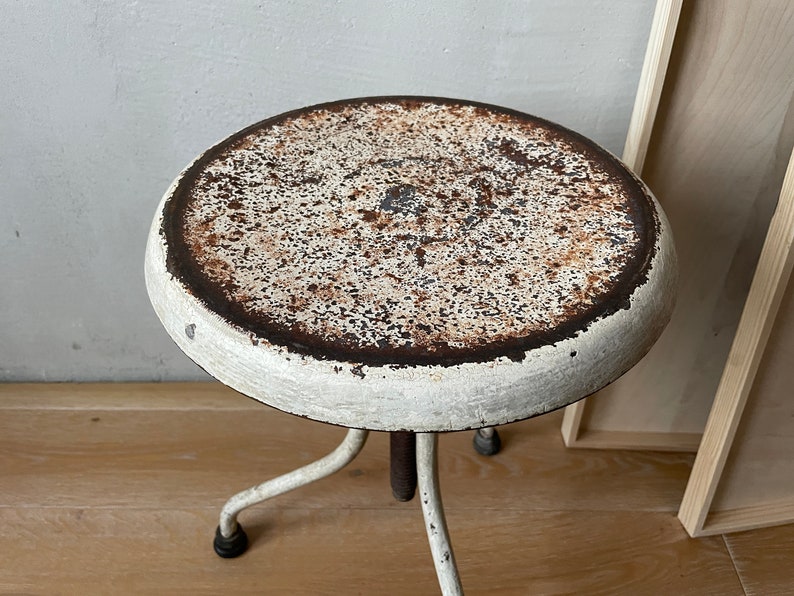 May include: White metal stool with a round, rusty top and three legs. The stool is slightly worn and has a vintage look.