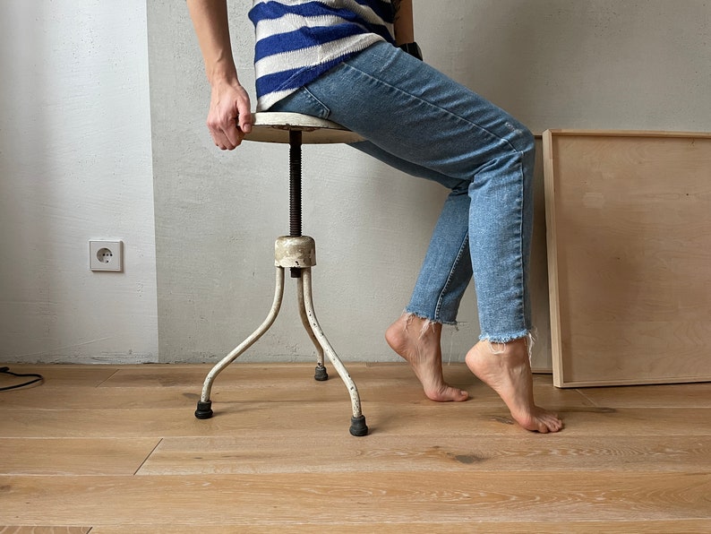 May include: A white metal adjustable stool with three legs. The stool has a round seat and a screw mechanism to adjust the height. The stool is in a room with wood floors.