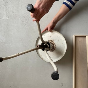 May include: White metal stool with three legs and black rubber feet. The stool is upside down and the legs are extended. The stool is being held by a person's hand.