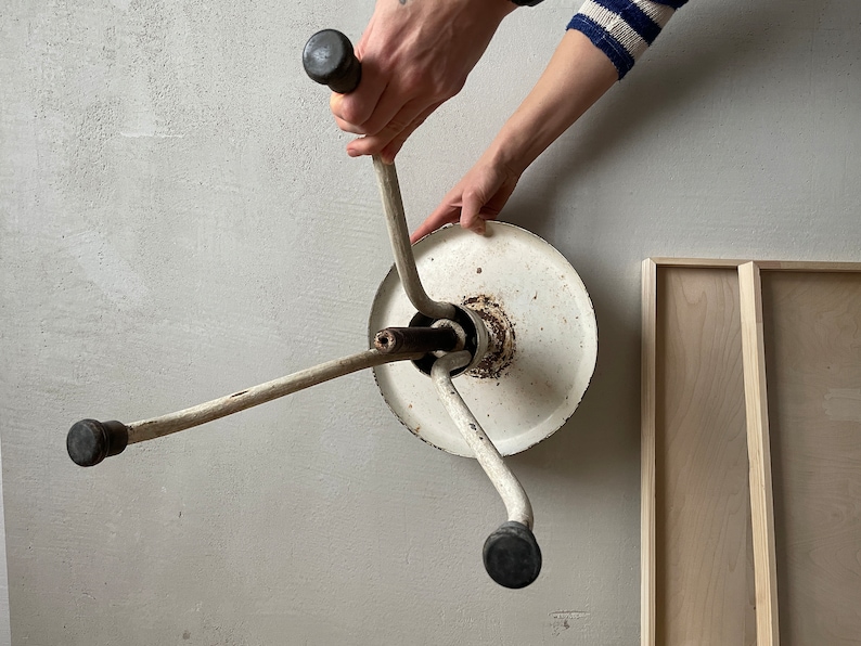 May include: White metal stool with three legs and black rubber feet. The stool is upside down and the legs are extended. The stool is being held by a person's hand.