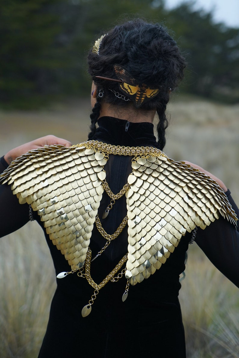 May include: A person wearing a black outfit with a gold chainmail collar and matching shoulder pieces. The shoulder pieces are shaped like wings and are made of small, overlapping scales. The person is also wearing a hairpiece with a butterfly design.