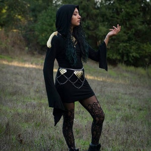May include: A person wearing a black hooded dress with gold chainmail accents and black lace tights. The person is standing in a grassy field with trees in the background.