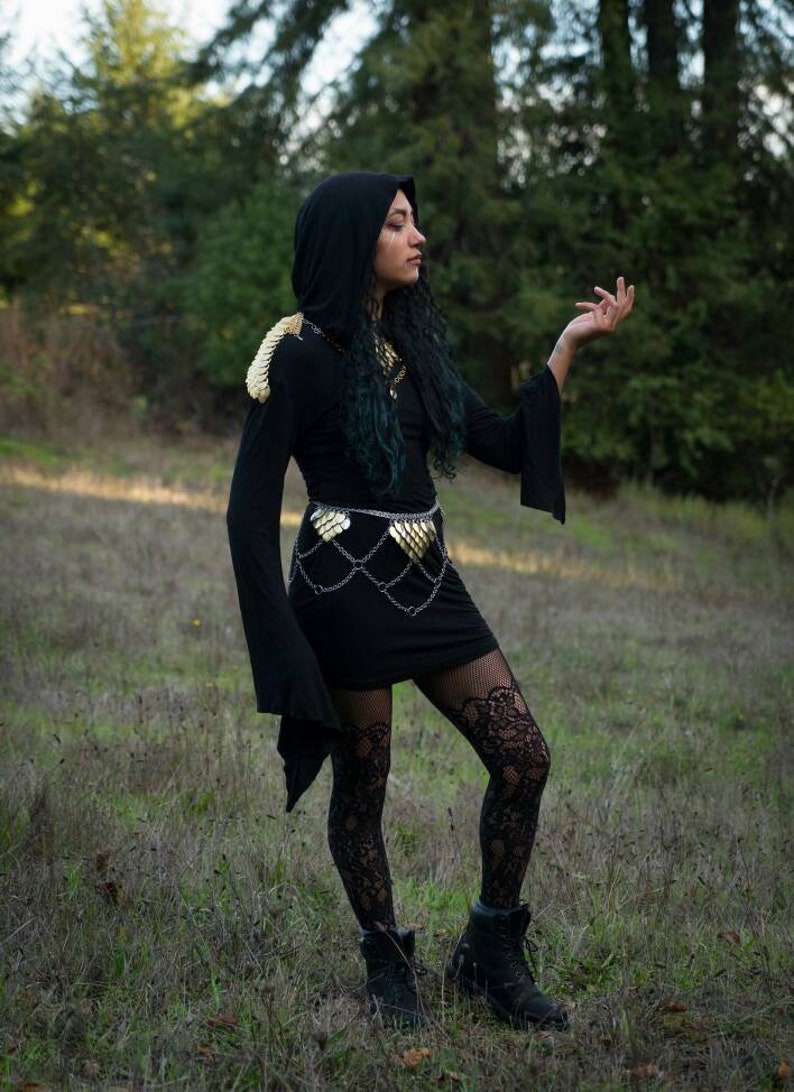 May include: A person wearing a black hooded dress with gold chainmail accents and black lace tights. The person is standing in a grassy field with trees in the background.