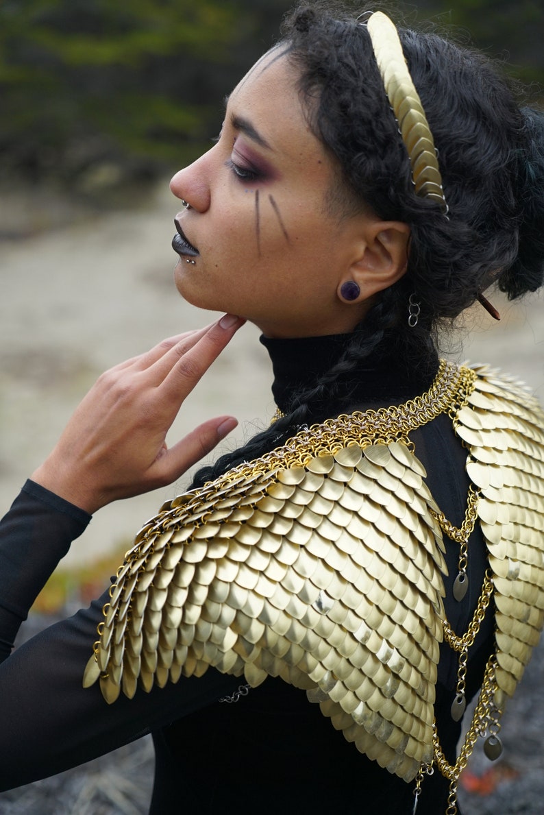 May include: A person wearing a black mock neck shirt with a gold chainmail collar and shoulder piece. The chainmail is made of small, overlapping scales.