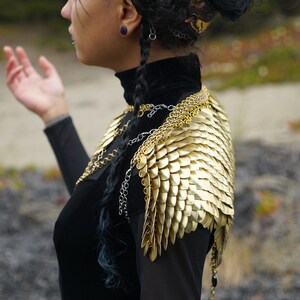 May include: A woman wearing a black turtleneck top with a gold chainmail shoulder piece and a gold headband.
