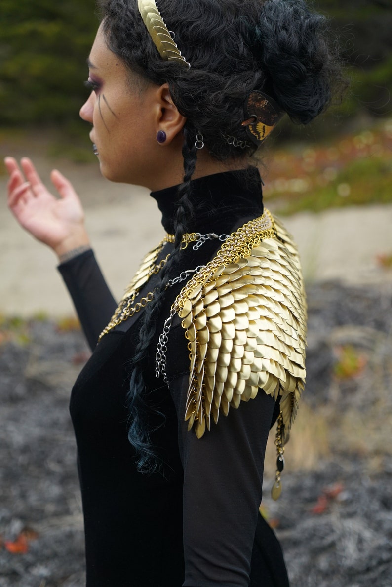 May include: A woman wearing a black turtleneck top with a gold chainmail shoulder piece and a gold headband.