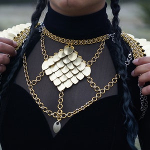 May include: A gold chainmail necklace with a large, heart-shaped pendant made of overlapping scales. The necklace is layered with several other chains, some silver and some gold.