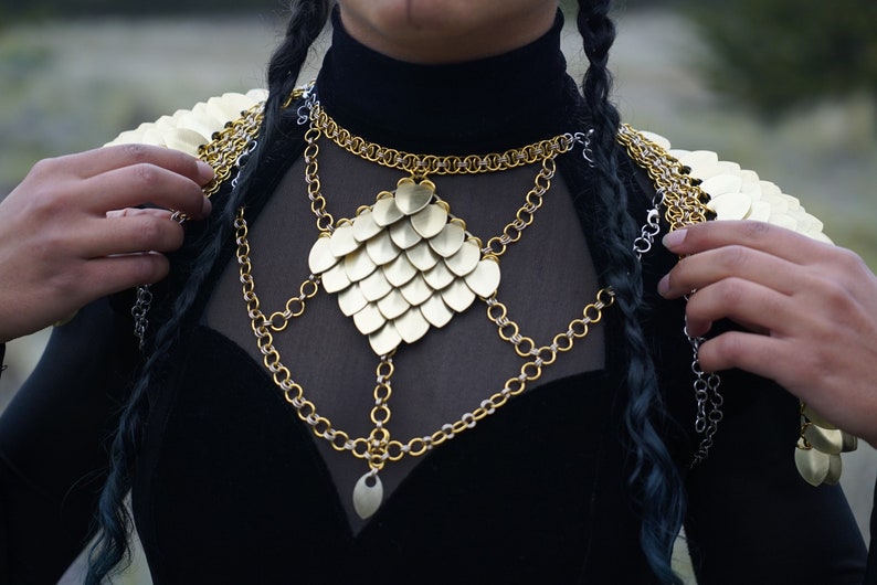 May include: A gold chainmail necklace with a large, heart-shaped pendant made of overlapping scales. The necklace is layered with several other chains, some silver and some gold.