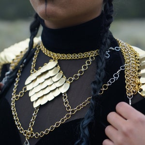 May include: A gold chainmail necklace with a large, heart-shaped pendant made of overlapping gold scales. The necklace is layered over a black shirt and worn with a matching gold chainmail shoulder piece.