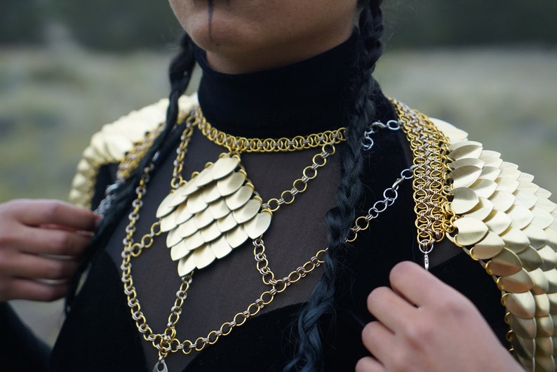 May include: A gold chainmail necklace with a large, heart-shaped pendant made of overlapping gold scales. The necklace is layered over a black shirt and worn with a matching gold chainmail shoulder piece.