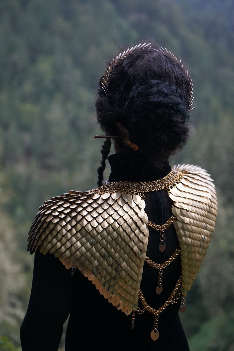 May include: A person wearing a black top with a gold chainmail shoulder piece and a gold chain with pendant charms. The person is wearing a crown of small metal spikes.