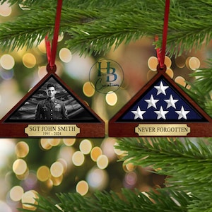 May include: Two wooden triangle-shaped ornaments with a red ribbon. The first ornament features a black and white photo of a man in military uniform with the text "SGT JOHN SMITH 1995 - 2024". The second ornament features a folded American flag with the text "NEVER FORGOTTEN".