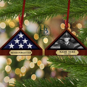 May include: Two wooden triangle-shaped ornaments with a folded American flag design. One ornament has a photo of a person in a military uniform and the text "NAME HERE 1944-2023". The other ornament has the text "NEVER FORGOTTEN".