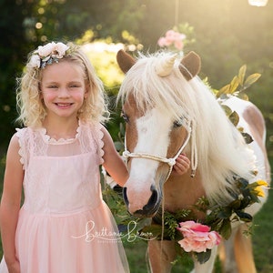 May include: A young girl in a pink dress with a flower crown is standing next to a white and brown pony with a unicorn horn. The pony is wearing a floral garland around its neck.