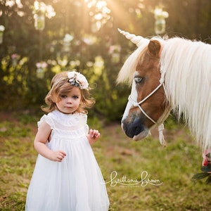 May include: A young girl in a white dress and flower crown looks at a white pony with a unicorn horn. The pony is wearing a floral garland around its neck.