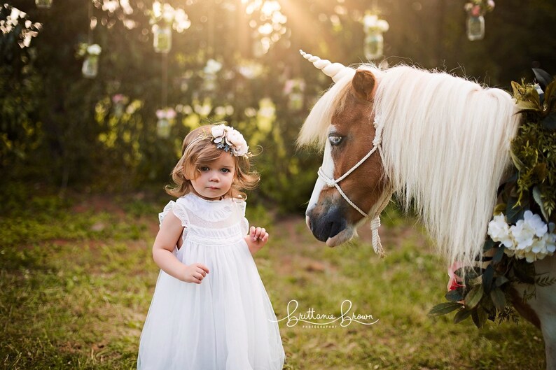 May include: A young girl in a white dress and flower crown looks at a white pony with a unicorn horn. The pony is wearing a floral garland around its neck.