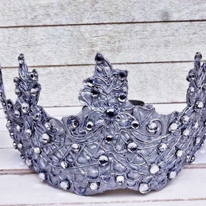 May include: A silver crown with a delicate floral design and rhinestones. The crown is made of metal and has a vintage look.