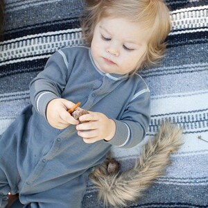 May include: A young child wearing a gray onesie with a white trim, lying on a striped blanket. The child is holding a small brown object in their hands.
