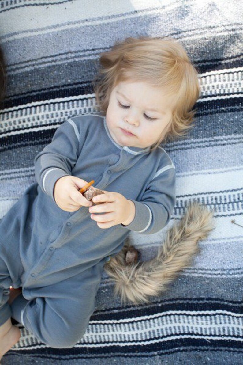 May include: A young child wearing a gray onesie with a white trim, lying on a striped blanket. The child is holding a small brown object in their hands.