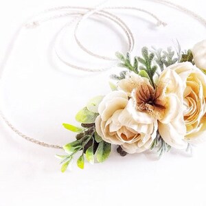 May include: A delicate floral crown with white and cream flowers and green leaves. The crown is made with a natural twine base.