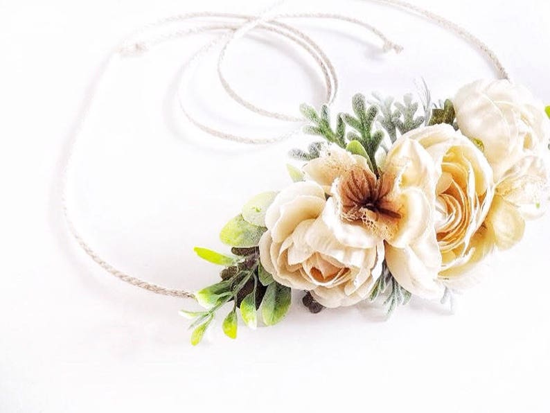 May include: A delicate floral crown with white and cream flowers and green leaves. The crown is made with a natural twine base.