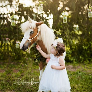 May include: A young girl in a white dress and flower crown reaches out to touch a brown and white pony with a unicorn horn. The pony is standing in a grassy field with a green tree in the background.