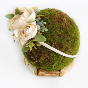 May include: A white headband with a floral arrangement of cream-colored flowers and green leaves. The headband is displayed on a bed of green moss.