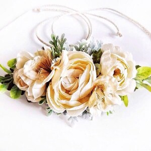 May include: A cream-colored floral crown with a simple, rustic rope base. The crown features three large, delicate flowers with a lace-like texture and small green leaves.