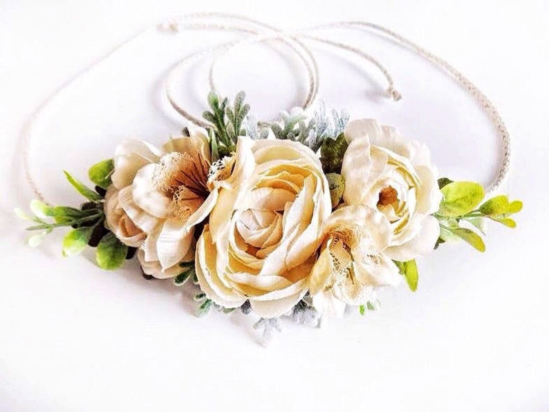 May include: A cream-colored floral crown with a simple, rustic rope base. The crown features three large, delicate flowers with a lace-like texture and small green leaves.