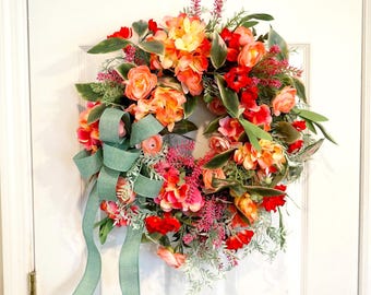 May include: A circular floral wreath featuring a mix of orange, red, and pink artificial flowers, green foliage, and small purple sprigs. A sage green fabric ribbon with long, flowing tails is tied to the lower left side of the wreath.