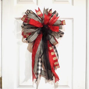 May include: A large decorative bow with multiple ribbons in red, black, and tan. The bow features a mix of solid and checkered patterns, creating a festive and eye-catching design. It is likely used for door decor or gift wrapping.