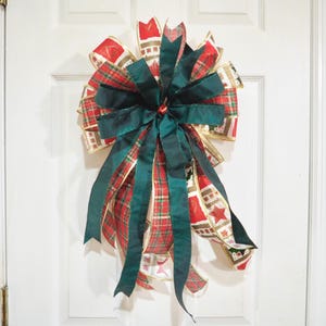 May include: A large Christmas bow with a layered design. The bow features red plaid and gold ribbon with festive patterns, accented by a deep green bow and streamers. The bow is attached to a white door.
