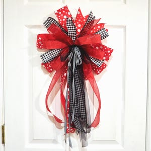 May include: A decorative bow featuring red, black, and white ribbons. The bow has a layered design with polka dot, houndstooth, and solid ribbons. Long streamers hang from the center, adding to the festive appearance. The bow is likely for seasonal or holiday decor.