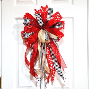 May include: A decorative door bow featuring red, black, and white ribbons. The bow has a layered design with polka dots, houndstooth, and solid red patterns. Gold and black-and-white striped ribbons hang down.